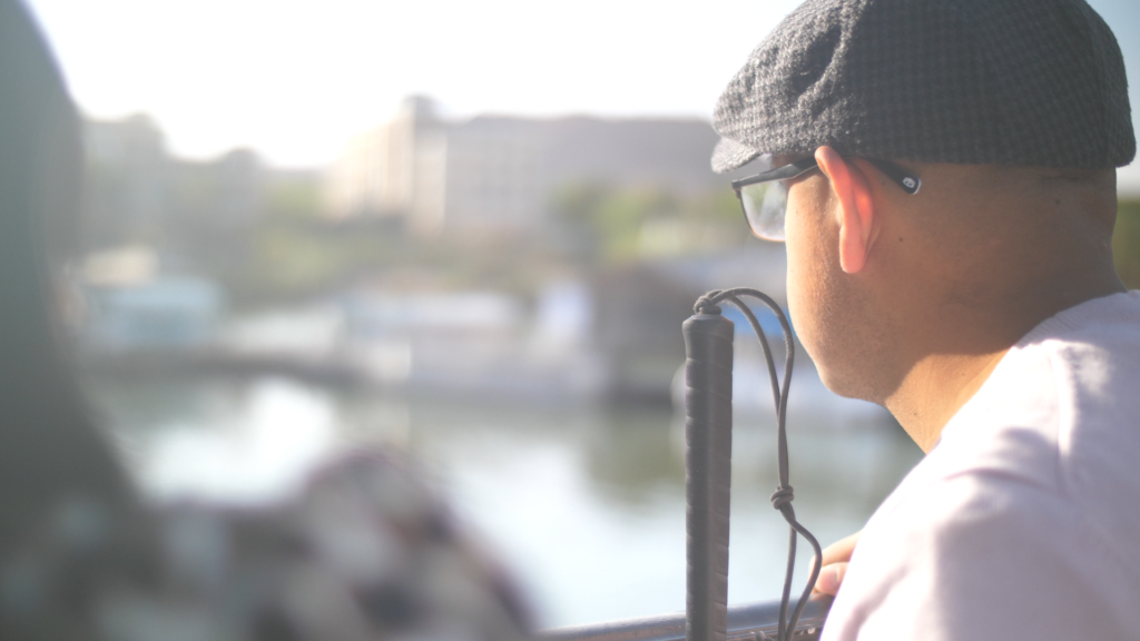 Still image: A man with glasses, gray beret, and light-colored shirt looks at an out-of-focus, daytime scenery. He has his back turned from the camera.